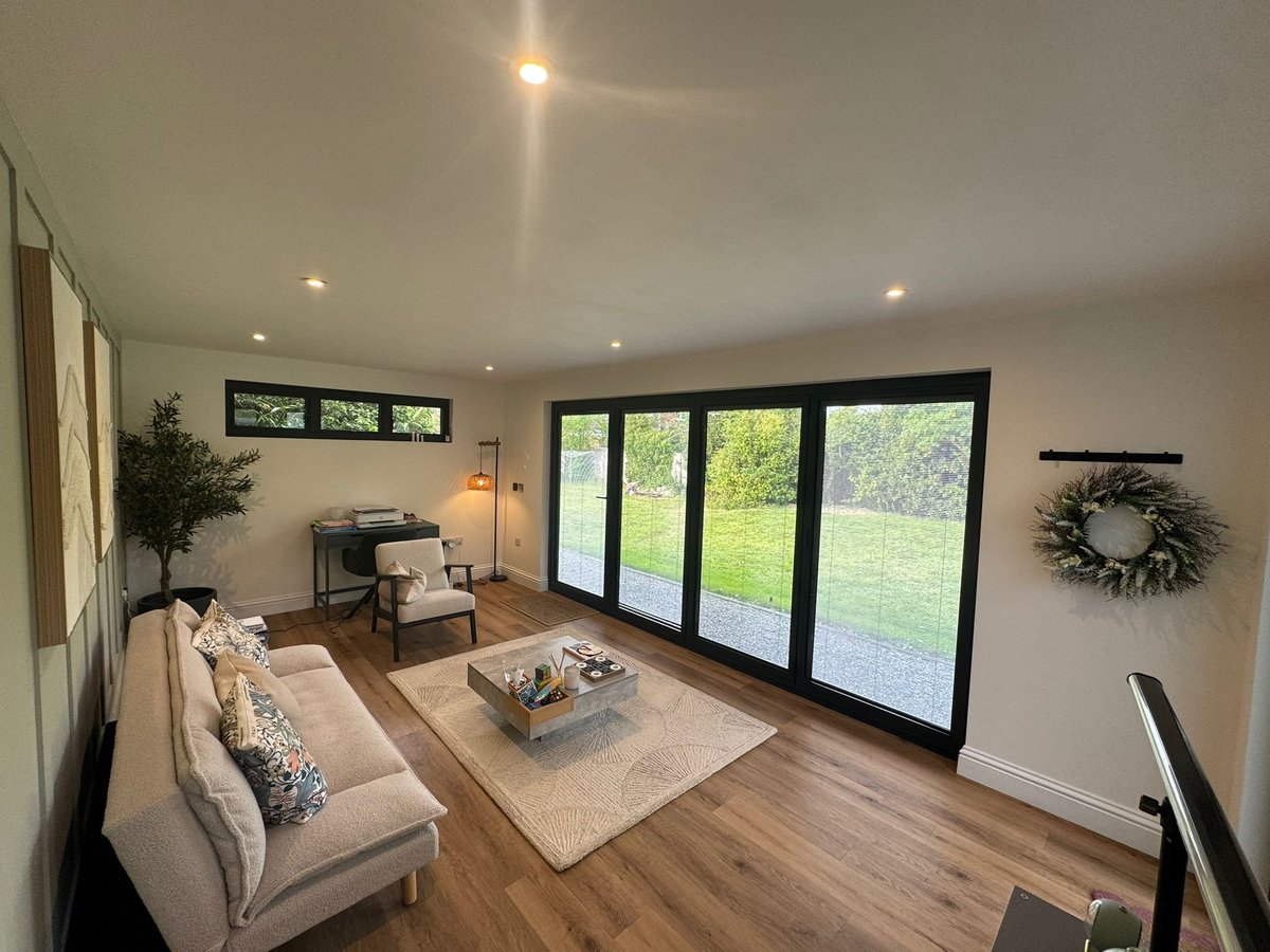 Modern living room with beige sectional sofa, glass doors overlooking green lawn, recessed lighting, and wooden flooring