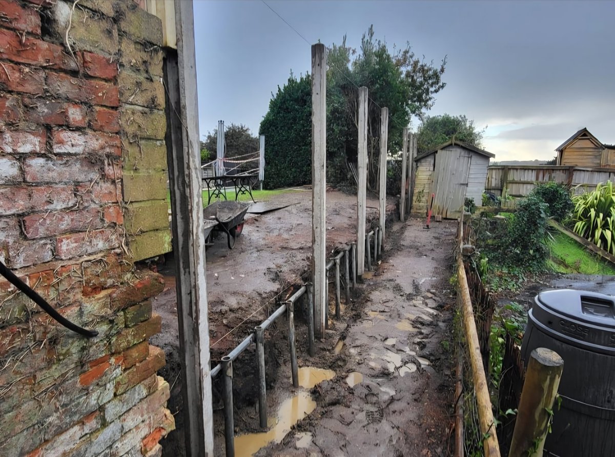 Muddy rural backyard garden with wooden fence posts, brick buildings, sheds, plants, and water barrel on cloudy day