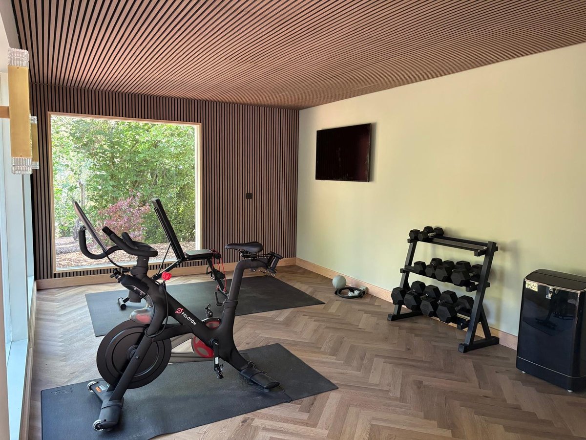 Modern home gym with stationary bike, dumbbells on rack, wooden ceiling, and large window overlooking garden