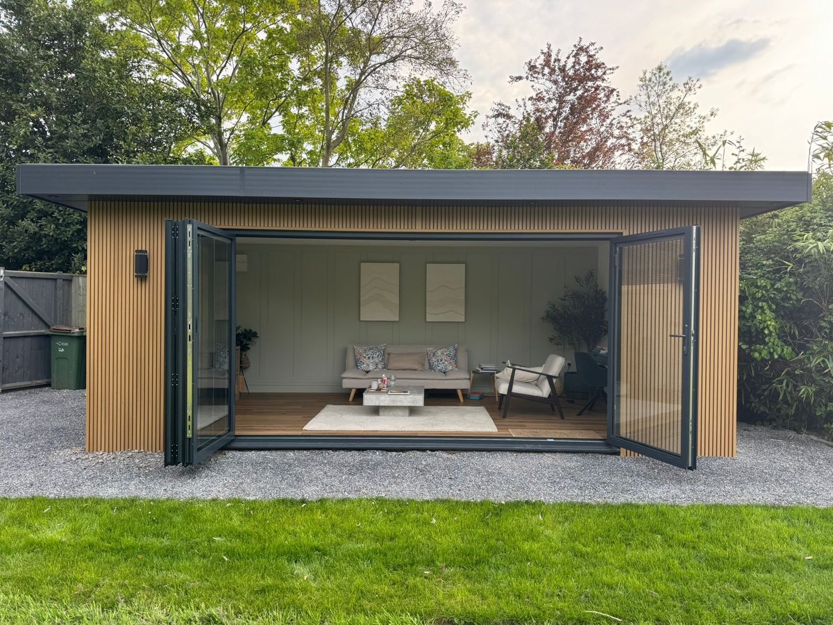 Modern garden studio with wooden slat exterior, open bifold doors revealing minimalist interior with seating and daybed, surrounded by grass and trees