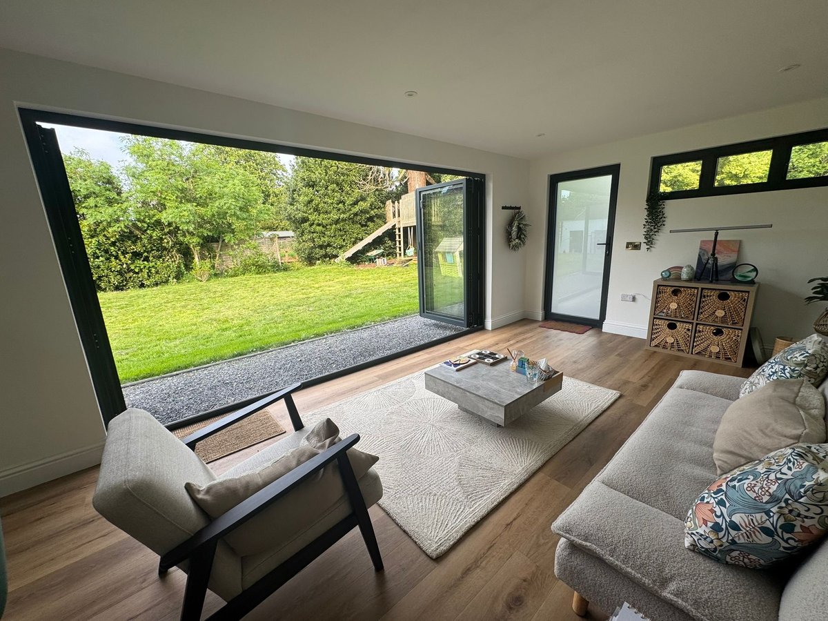 Modern living room with large glass doors opening to a green garden, featuring neutral furnishings and wooden flooring