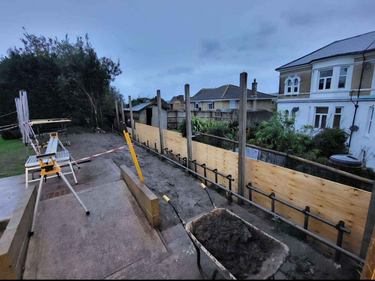 Residential backyard under construction with wooden fencing, concrete foundation, and building materials visible, adjacent to a light blue house.