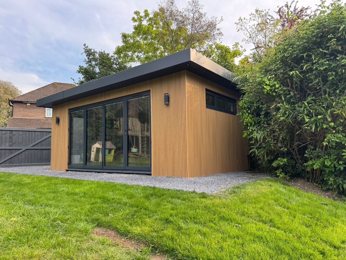 Modern garden studio with wood cladding, black roof, and large windows set on a gravel base in a green lawn