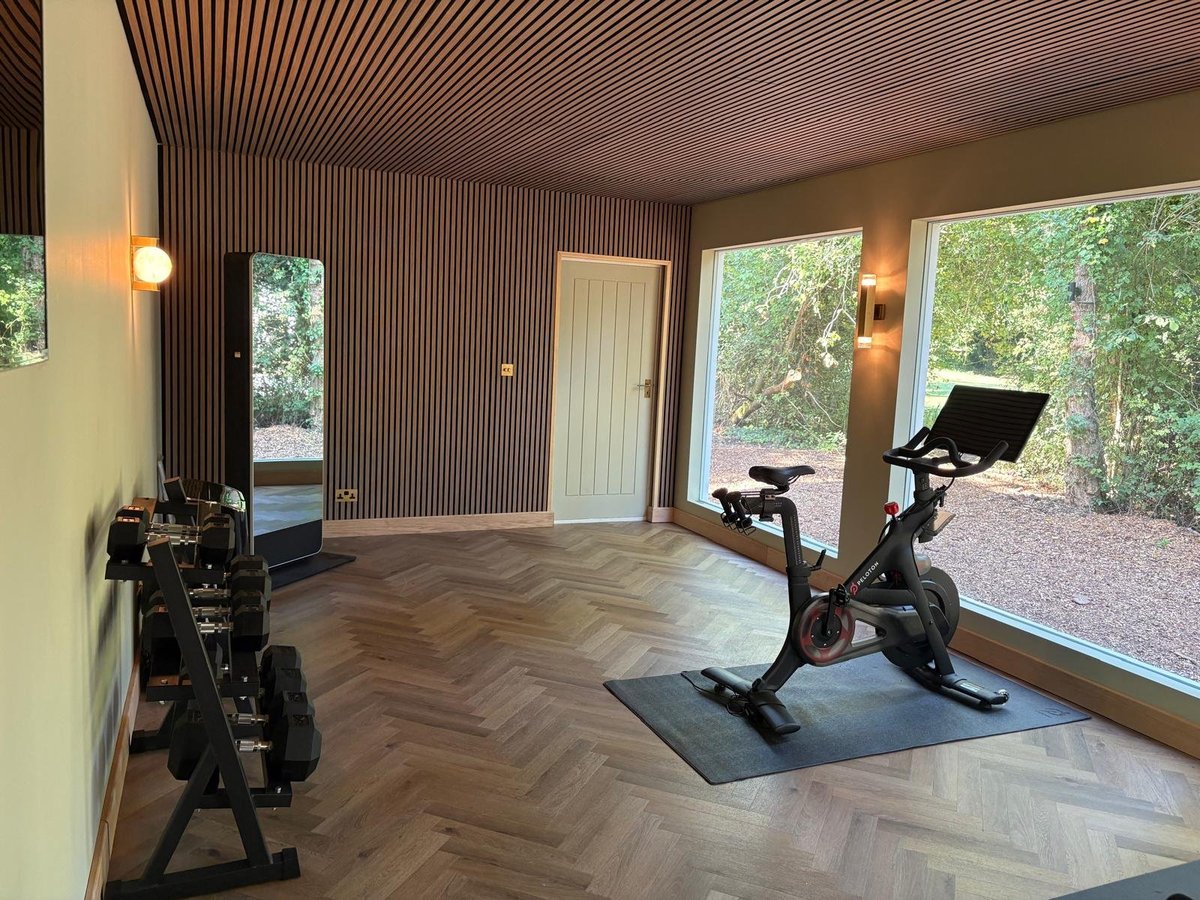 Modern home gym with two stationary bikes on wood flooring, large windows overlooking a garden, and minimalist decor