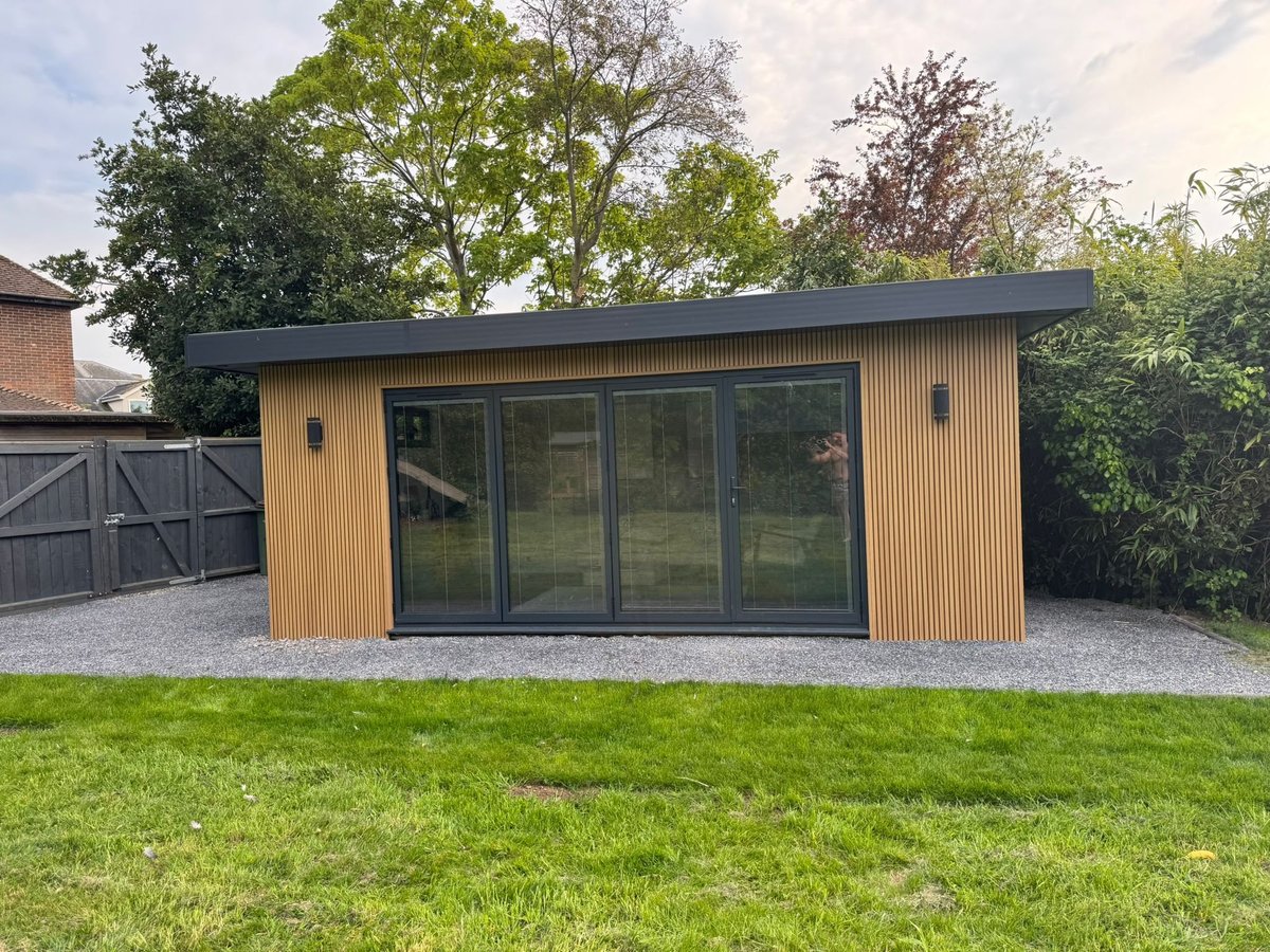 Modern garden studio with wooden cladding, dark roof, and large glass sliding doors on a gravel patio surrounded by grass and trees